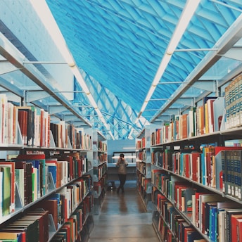 A modern library interior featuring long aisles lined with bookshelves filled with various colorful books. The scene is framed by a high, geometric ceiling with a striking blue pattern. In the center of the aisle, a person is taking a photograph, adding a touch of dynamic presence to the serene environment.