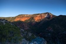 A striking landscape featuring a rugged mountain with steep cliffs. The warm, reddish hues of the rock formations stand out against the clear blue sky. Dense green vegetation covers parts of the slopes, adding contrast to the rugged terrain.