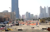 A construction site with several workers wearing blue uniforms is visible in the foreground. Heavy machinery and equipment are present, including a bulldozer. The site is surrounded by safety barriers and orange netting. In the background, a city skyline of modern skyscrapers and tall buildings can be seen.