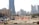 A construction site with several workers wearing blue uniforms is visible in the foreground. Heavy machinery and equipment are present, including a bulldozer. The site is surrounded by safety barriers and orange netting. In the background, a city skyline of modern skyscrapers and tall buildings can be seen.
