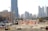 A construction site with several workers wearing blue uniforms is visible in the foreground. Heavy machinery and equipment are present, including a bulldozer. The site is surrounded by safety barriers and orange netting. In the background, a city skyline of modern skyscrapers and tall buildings can be seen.