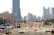 A construction site with several workers wearing blue uniforms is visible in the foreground. Heavy machinery and equipment are present, including a bulldozer. The site is surrounded by safety barriers and orange netting. In the background, a city skyline of modern skyscrapers and tall buildings can be seen.