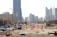 A construction site with several workers wearing blue uniforms is visible in the foreground. Heavy machinery and equipment are present, including a bulldozer. The site is surrounded by safety barriers and orange netting. In the background, a city skyline of modern skyscrapers and tall buildings can be seen.