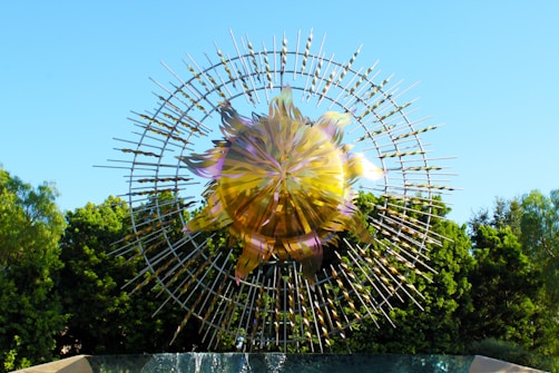 A large, intricate sculpture resembling a radiant sun is positioned against a clear blue sky. The sculpture consists of a shiny, metallic circular center with numerous elongated spikes extending outward, creating a dynamic and energetic pattern. Lush green trees form the backdrop, enhancing the sculpture’s reflective qualities.