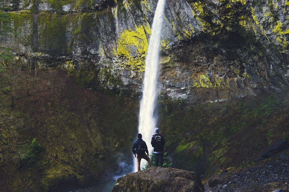 two persons standing on rock near waterfall at daytime