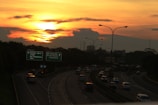 A highway with multiple lanes features cars heading in various directions under a vibrant sunset. The sky is a mix of orange and yellow hues, with scattered clouds. Road signs indicate destinations like Bambu Apus, Ceger, Jagorawi, and more. Streetlights line the road.