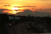 A highway with multiple lanes features cars heading in various directions under a vibrant sunset. The sky is a mix of orange and yellow hues, with scattered clouds. Road signs indicate destinations like Bambu Apus, Ceger, Jagorawi, and more. Streetlights line the road.