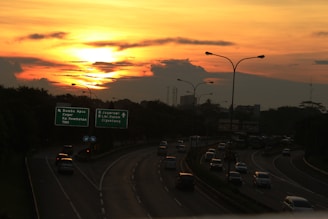 A highway with multiple lanes features cars heading in various directions under a vibrant sunset. The sky is a mix of orange and yellow hues, with scattered clouds. Road signs indicate destinations like Bambu Apus, Ceger, Jagorawi, and more. Streetlights line the road.