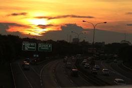 A highway with multiple lanes features cars heading in various directions under a vibrant sunset. The sky is a mix of orange and yellow hues, with scattered clouds. Road signs indicate destinations like Bambu Apus, Ceger, Jagorawi, and more. Streetlights line the road.