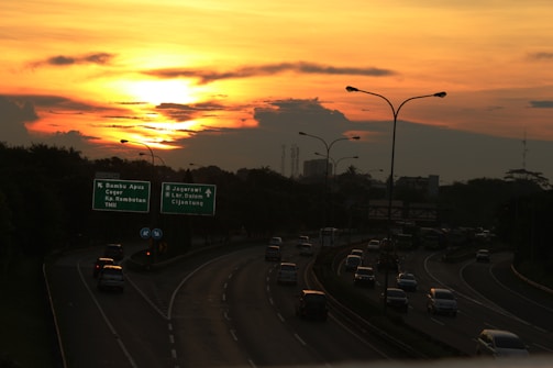 A highway with multiple lanes features cars heading in various directions under a vibrant sunset. The sky is a mix of orange and yellow hues, with scattered clouds. Road signs indicate destinations like Bambu Apus, Ceger, Jagorawi, and more. Streetlights line the road.