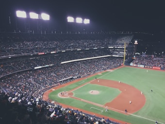 A nighttime baseball game in a large, brightly lit stadium with numerous spectators in the stands. Players are positioned on the field as the game is underway.