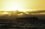 A large LNG ship is silhouetted against the dramatic backdrop of a sunset over the ocean. The sun is partially obscured by thick, low-lying clouds, casting a golden glow across the sky and creating reflections on the water.