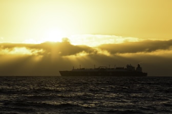 A large LNG ship is silhouetted against the dramatic backdrop of a sunset over the ocean. The sun is partially obscured by thick, low-lying clouds, casting a golden glow across the sky and creating reflections on the water.