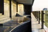 A modern balcony or corridor area with a curved granite handrail and glass panels supported by metal fixtures. The warm light casts subtle shadows and reflections on the granite surface and walls, creating a serene and minimalistic architectural space.