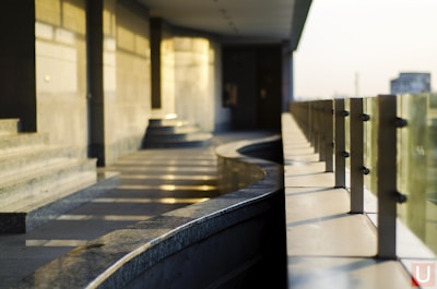 A modern balcony or corridor area with a curved granite handrail and glass panels supported by metal fixtures. The warm light casts subtle shadows and reflections on the granite surface and walls, creating a serene and minimalistic architectural space.