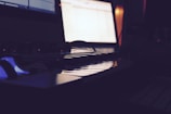 A dimly lit room features a digital keyboard or synthesizer in the foreground, with a computer screen glowing in the background, displaying some open software or application.