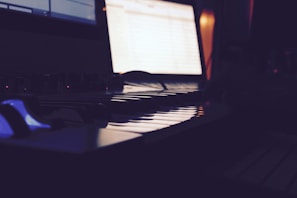 A dimly lit room features a digital keyboard or synthesizer in the foreground, with a computer screen glowing in the background, displaying some open software or application.