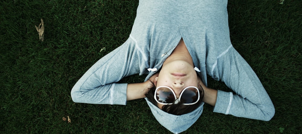 A person in dark glasses and a gray hoodie lying relaxed on green grass