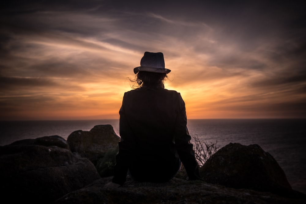 Silhouette eines Mannes, der auf einem Stein sitzt und auf ein Gewässer schaut