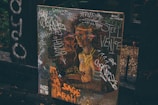 A painting or poster depicting a classical-style figure, covered in colorful graffiti and scribbles. The background includes dark wooden surfaces and foliage, adding an urban or alleyway vibe. The presence of words and drawings suggests a blend of art and street culture.