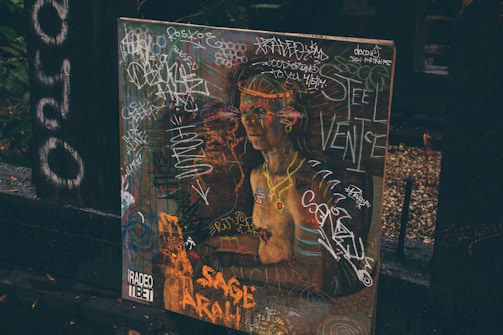 A painting or poster depicting a classical-style figure, covered in colorful graffiti and scribbles. The background includes dark wooden surfaces and foliage, adding an urban or alleyway vibe. The presence of words and drawings suggests a blend of art and street culture.