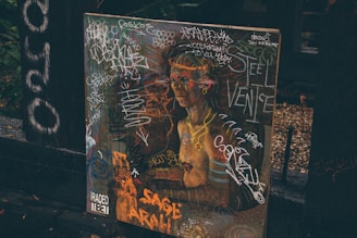 A painting or poster depicting a classical-style figure, covered in colorful graffiti and scribbles. The background includes dark wooden surfaces and foliage, adding an urban or alleyway vibe. The presence of words and drawings suggests a blend of art and street culture.
