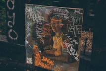 A painting or poster depicting a classical-style figure, covered in colorful graffiti and scribbles. The background includes dark wooden surfaces and foliage, adding an urban or alleyway vibe. The presence of words and drawings suggests a blend of art and street culture.