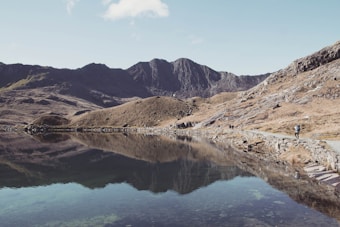 A serene mountain landscape featuring a still lake that perfectly mirrors the surrounding rugged terrain and sky. The shoreline stretches along the right side of the image, where a lone hiker walks along a stone path bordered by rough, rocky hills. The distant mountains are bathed in soft sunlight, highlighting their jagged peaks against a clear blue sky.