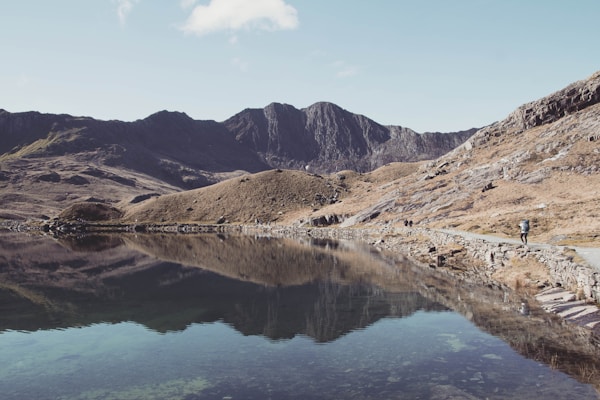 A serene mountain landscape featuring a still lake that perfectly mirrors the surrounding rugged terrain and sky. The shoreline stretches along the right side of the image, where a lone hiker walks along a stone path bordered by rough, rocky hills. The distant mountains are bathed in soft sunlight, highlighting their jagged peaks against a clear blue sky.