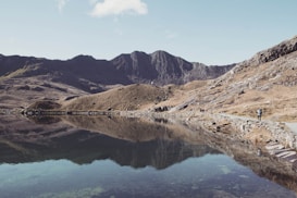 A serene mountain landscape featuring a still lake that perfectly mirrors the surrounding rugged terrain and sky. The shoreline stretches along the right side of the image, where a lone hiker walks along a stone path bordered by rough, rocky hills. The distant mountains are bathed in soft sunlight, highlighting their jagged peaks against a clear blue sky.