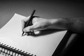 A hand holding a pen poised over a spiral-bound notebook. The scene is in black and white, highlighting the textures of the skin and paper. The notebook is open to a blank page, and the pen is being held gently between the fingers, suggesting the start of a drawing or note.