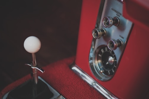 Close-up view of a vintage car dashboard featuring three metallic control knobs labeled defroster, fan, and temp, alongside a speedometer or clock. The design has a classic aesthetic with a red interior and an extended white gear shift knob on the left.