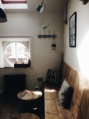 A cozy interior space featuring a wooden bench with cushions along a wall. Above the bench, a green light fixture and a black-and-white framed picture are mounted. A small round window with several items on the sill lets in natural light. The floor is wooden, and there are two small round wooden tables near the bench. A coat rack with hooks and a hanging plant are also visible on the wall.