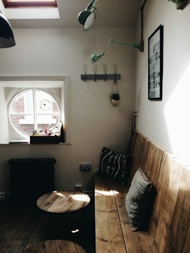 A cozy interior space featuring a wooden bench with cushions along a wall. Above the bench, a green light fixture and a black-and-white framed picture are mounted. A small round window with several items on the sill lets in natural light. The floor is wooden, and there are two small round wooden tables near the bench. A coat rack with hooks and a hanging plant are also visible on the wall.