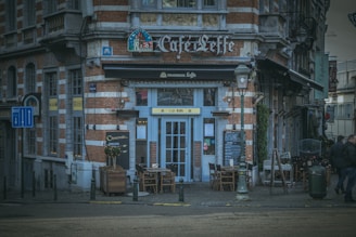 A quaint street-side café with a classic brick facade and blue accents. The front is adorned with signage, a menu board, and tables arranged outside. Street lamps and pedestrians enhance the European city atmosphere.