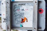 A weathered wooden frame surrounds a metal sign covered with various stickers and graffiti. The stickers feature different logos, symbols, and text, some with bold designs and bright colors. The metal surface appears worn and slightly rusted, adding to the urban and gritty feel.