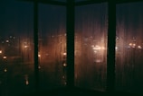 Rain streaking down a foggy window overlooking a quiet city street at night.