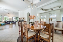 A spacious and well-lit open-plan living area features a wooden dining table with matching chairs in the foreground. The room has white walls with large windows allowing natural light to pour in. In the background, there's a cozy seating area with sofas and potted plants. The floors are tiled, and modern light fixtures hang from the ceiling.