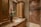 A luxurious bathroom with a detailed marble countertop and a bronze faucet. A decorative flower arrangement in a vase adds elegance. Wooden cabinets with glass doors offer storage, complemented by overhead lighting. The walk-in shower features large, diagonally laid brown tiles and a glass door.