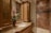 A luxurious bathroom with a detailed marble countertop and a bronze faucet. A decorative flower arrangement in a vase adds elegance. Wooden cabinets with glass doors offer storage, complemented by overhead lighting. The walk-in shower features large, diagonally laid brown tiles and a glass door.