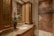 A luxurious bathroom with a detailed marble countertop and a bronze faucet. A decorative flower arrangement in a vase adds elegance. Wooden cabinets with glass doors offer storage, complemented by overhead lighting. The walk-in shower features large, diagonally laid brown tiles and a glass door.