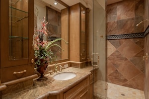 A luxurious bathroom with a detailed marble countertop and a bronze faucet. A decorative flower arrangement in a vase adds elegance. Wooden cabinets with glass doors offer storage, complemented by overhead lighting. The walk-in shower features large, diagonally laid brown tiles and a glass door.