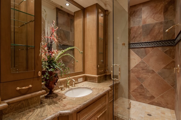 A luxurious bathroom with a detailed marble countertop and a bronze faucet. A decorative flower arrangement in a vase adds elegance. Wooden cabinets with glass doors offer storage, complemented by overhead lighting. The walk-in shower features large, diagonally laid brown tiles and a glass door.