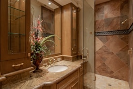 A luxurious bathroom with a detailed marble countertop and a bronze faucet. A decorative flower arrangement in a vase adds elegance. Wooden cabinets with glass doors offer storage, complemented by overhead lighting. The walk-in shower features large, diagonally laid brown tiles and a glass door.