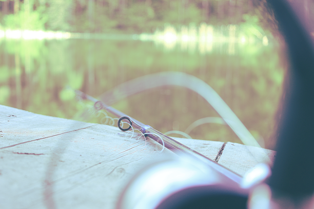 A close-up of a fishing rod and reel resting on a wooden dock with a calm lake in the background at sunrise.