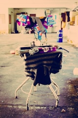 A makeshift sculpture created from assorted items such as cans, cloth, and tape is positioned on a worn metal chair. The background features an industrial setting with a large, open space that appears unused or abandoned. The floor is scattered with debris, adding to the overall sense of neglect.