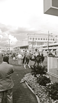 A black and white image depicting a horse race event. The foreground shows people gathered around a track preparing a horse with the number 3 on its saddle. The track is surrounded by a low railing, with flowers planted along the edge. In the background, there is a large building with multiple balconies filled with people watching. The atmosphere appears busy and lively.