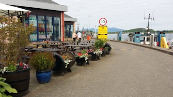 A waterfront area with outdoor seating featuring picnic tables and decorative barrel planters filled with colorful flowers. A small building with large windows is positioned to the left, and there is a road with traffic signs, including a speed limit sign. A fishing boat is docked by the water, with hills visible in the background.