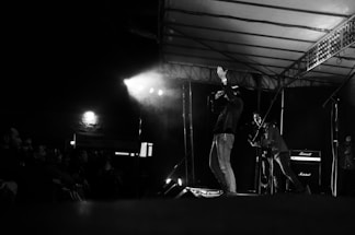A black and white image captures a live music performance on stage. A singer holds a microphone, gesturing with one hand, while another musician plays a guitar. The stage is dimly lit with spotlights creating dramatic shadows. Amplifiers are visible in the background, and an audience watches from the side.