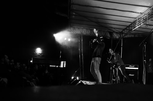 A black and white image captures a live music performance on stage. A singer holds a microphone, gesturing with one hand, while another musician plays a guitar. The stage is dimly lit with spotlights creating dramatic shadows. Amplifiers are visible in the background, and an audience watches from the side.
